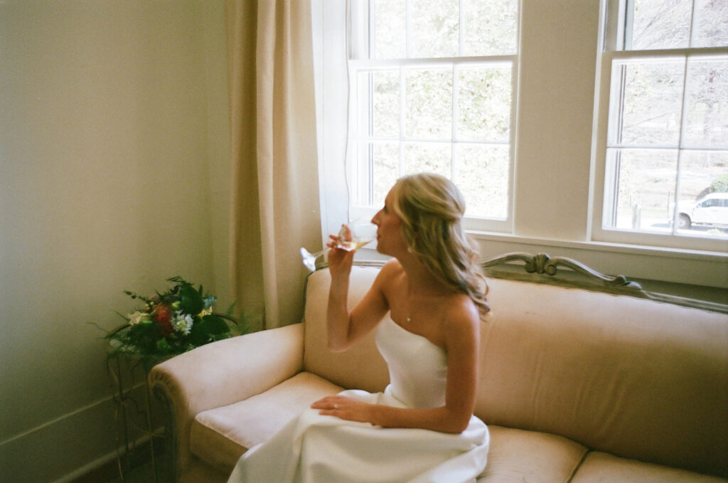 film shot of bride drinking a glass of champagne on a white couch with florals to the side