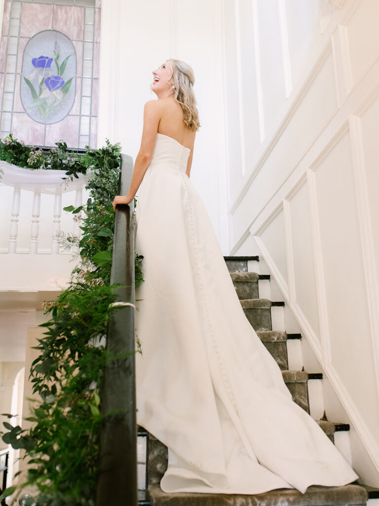 bridal gown, white on the stairs with greenery on the handle. bride looking up with sweeping train coming down the steps.