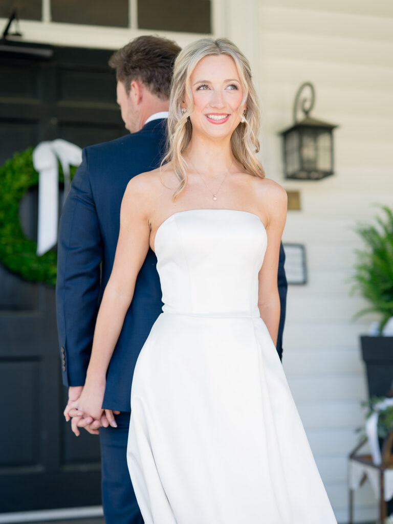 bride and groom on the front porch holding hands looking away from each other. smiling big with white strapless dress.
