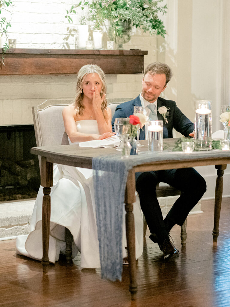 bride and groom sitting at a table. bride wiping tears away as groom holds her hand during reception at payne corley house