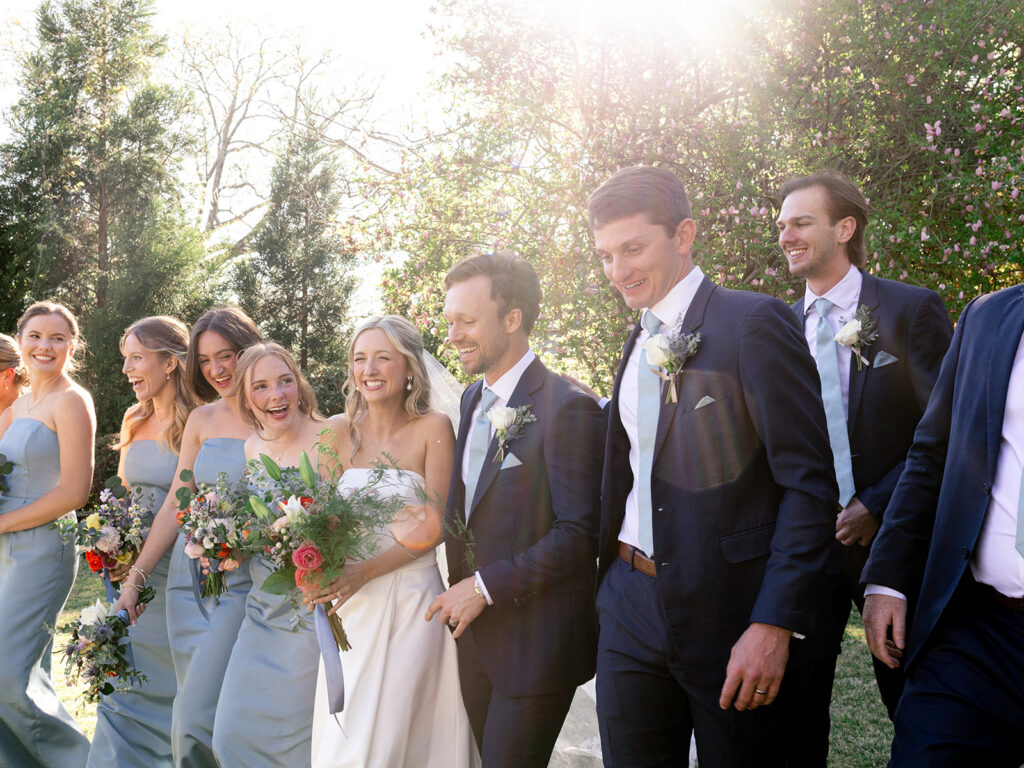 group of bridal party walking away from camera. smiling big and laughing with lens flare of the sunny day